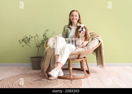 Jeune femme et adorable cavalier King Charles spaniel assis sur un fauteuil près du mur vert à la maison Banque D'Images