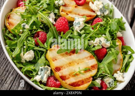 salade de poires grillées avec fromage bleu, roquette et framboises fraîches mûres dans un bol blanc sur une table en bois rustique, vue d'angle hollandais, gros plan Banque D'Images