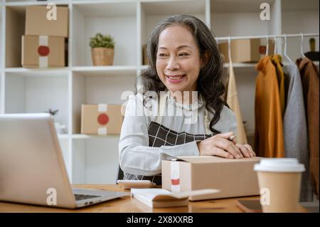 Un heureux vendeur en ligne de vêtements féminins asiatiques matures ou couturière vérifie les commandes des clients sur son ordinateur, emballe les colis d'expédition, et travaille Banque D'Images