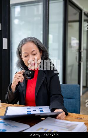 Une femme d'affaires asiatique mature confiante et satisfaite ou un patron féminin sirote un café tout en lisant le rapport d'affaires à son bureau. Banque D'Images