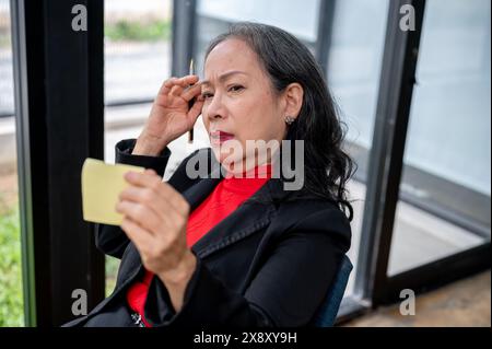 Une femme d'affaires asiatique senior regarde une note collante avec une expression insatisfaite, sentant un mal de tête et confus après avoir lu le texte sur le Banque D'Images