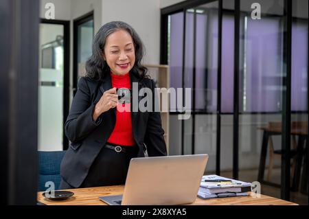 Une femme d'affaires asiatique mature et satisfaite ou un patron féminin se tient debout à son bureau et sirote un café tout en lisant des rapports sur son ordinateur portable dans le Banque D'Images