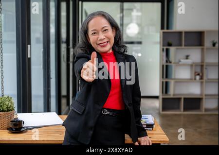 Une femme d'affaires asiatique mature confiante et attrayante dans un costume formel montre son pouce vers le haut et sourit à la caméra tout en se tenant debout dans le bureau. Banque D'Images