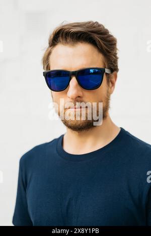 Portrait d'un homme debout avec confiance contre un mur blanc, portant des lunettes de soleil réfléchissantes et un t-shirt bleu, exsudant un comportement frais et composé. Banque D'Images