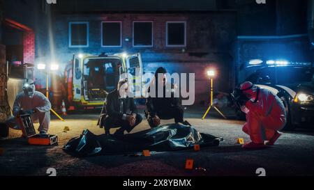 Équipe d'enquête sur la scène de crime travaillant sur un cas d'homicide la nuit. Spécialiste en médecine légale prenant professionnellement des photos de la victime morte dans un sac corporel. Détectives discutant des scénarios potentiels Banque D'Images