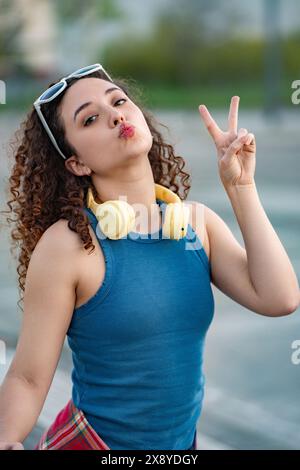 Jeune femme joyeuse avec les cheveux bouclés qui se plient les lèvres, faisant un signe de paix tout en portant des écouteurs à l'extérieur. Banque D'Images