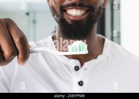 A la maison, homme afro-américain se brossant les dents dans la salle de bain. Porter une chemise blanche et porter une barbe, tenir une brosse à dents avec de la pâte, inaltérée Banque D'Images