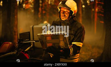 Portrait d'un beau pompier dans l'équipement de sécurité à l'aide d'un ordinateur portable robuste, rapport sur une situation avec un incendie dangereux de Wildland dans une forêt. Dure journée au travail pour un pompier volontaire. Banque D'Images