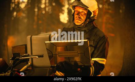 Portrait d'un beau pompier dans l'équipement de sécurité à l'aide d'un ordinateur portable robuste, rapport sur une situation avec un incendie dangereux de Wildland dans une forêt. Dure journée au travail pour un pompier volontaire courageux. Banque D'Images