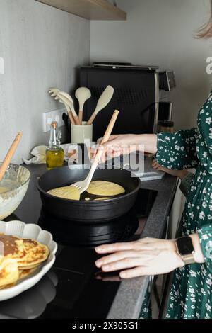 Main de femme tenant la spatule avec des crêpes dans la poêle à frire sur la cuisinière Banque D'Images