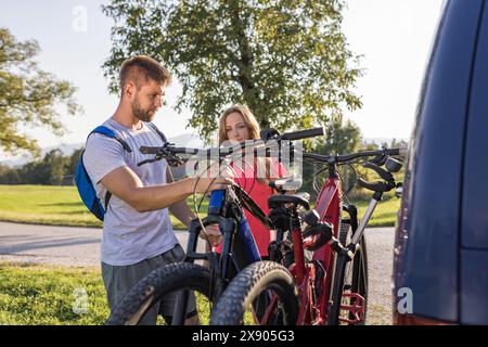 Le jeune homme décharge les vélos de montagne électriques, pour lui et sa petite amie, les soulevant de la galerie d'attelage du véhicule. Banque D'Images