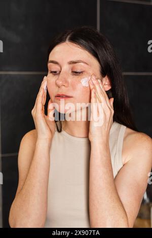 La jeune brune applique de la crème solaire sur son visage. Routine de soin de la peau. Concept de soin de la peau. Banque D'Images