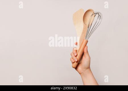 Main tenant une cuillère en bois, une spatule en bois et un fouet sur un fond beige Uni, copier l'espace Banque D'Images