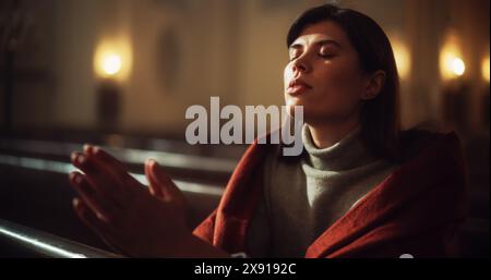 Jeune femme chrétienne est assise pieusement dans l'église Majestic, avec les mains pliées elle cherche la guidance de la foi et de la spiritualité tout en priant. Croyance religieuse en puissance et amour de Dieu. Prise de vue cinématographique Banque D'Images
