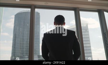 Homme d'affaires prospère se tient près de la fenêtre panoramique. Vue arrière de l'homme d'affaires réfléchi portant un costume debout dans son bureau, les mains dans les poches Banque D'Images