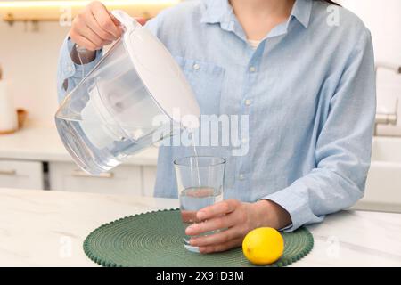 Femme versant de l'eau de la cruche filtrante dans le verre à la table en marbre blanc dans la cuisine, gros plan Banque D'Images