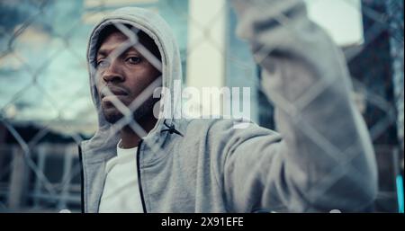Portrait d'un jeune homme noir attrayant portant un sweat à capuche, tenant une clôture, posant et regardant la caméra. Ambiance de séance photo commerciale pour une marque de streetwear jeunesse. Gros plan Banque D'Images