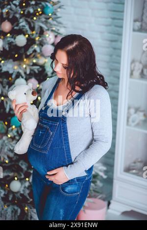 Femme enceinte en salopette en denim et pull gris tenant un ours en peluche devant un arbre de Noël décoré Banque D'Images