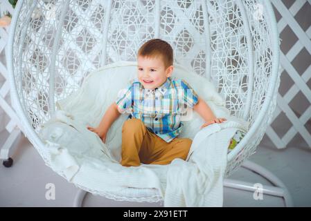 Un jeune garçon souriant portant une chemise à carreaux et un pantalon beige est assis dans un confortable fauteuil d'oeuf blanc à l'intérieur Banque D'Images