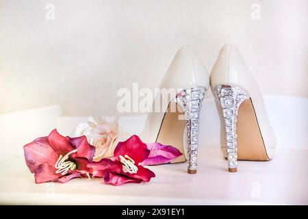 Talons hauts blancs fantaisie avec des détails de strass à côté de fleurs roses sur un banc blanc créant une atmosphère nuptiale élégante Banque D'Images