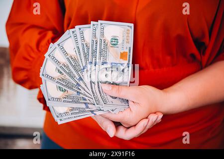 Personne tenant un éventail de billets de 100 dollars dans les deux mains portant une chemise rouge Banque D'Images