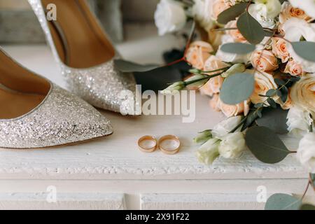 Élégantes chaussures à paillettes et anneaux de mariage placés près d'un bouquet de mariage de fleurs blanches et de roses pêche avec des feuilles d'eucalyptus sur une surface en bois Banque D'Images