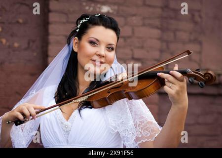 Mariée dans une robe blanche joue du violon à côté d'un mur de briques, avec un voile et accessoire de cheveux floral, exsudant une atmosphère paisible. Biélorussie, Minsk Banque D'Images