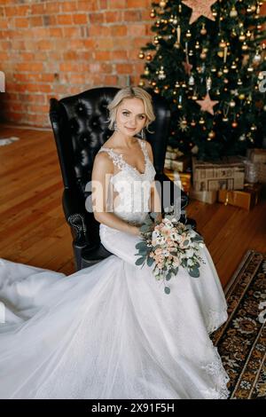 Mariée dans une robe de mariée tenant un bouquet, assise sur une chaise en cuir noir avec un arbre de Noël et des décorations derrière elle Banque D'Images