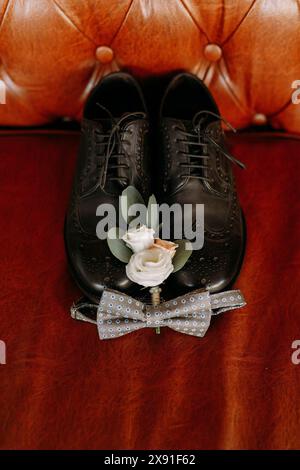 Chaussures de mariage en cuir noir avec une boutonnière florale blanche et un noeud papillon bleu placé sur un canapé en cuir marron Banque D'Images