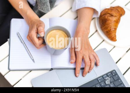 Une femme travaille sur un ordinateur portable, prend des notes et boit du café pendant une réunion matinale en ligne dans un café. Banque D'Images