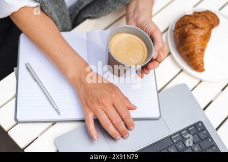 Une femme travaille sur un ordinateur portable, prend des notes et boit du café pendant une réunion matinale en ligne dans un café. Banque D'Images