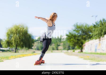 Une jeune femme en haut rayé et roller balance ludique sur un sentier ensoleillé dans le parc, montrant de la joie et un style de vie actif. Banque D'Images