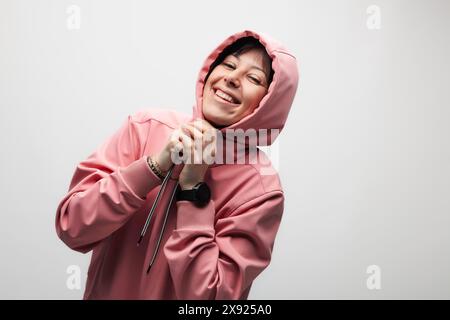 Une jeune femme joyeuse vêtue d'un sweat à capuche rose sourit largement, rayonnant de bonheur et de positivité sur un fond Uni. Banque D'Images