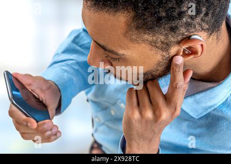 Jeune homme ajustant l'intensité du volume de l'aide auditive avec le smartphone. Digital Hearing-Adjustments avec le smartphone. 016561 067 Banque D'Images