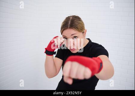 Combattante déterminée avec des bandes rouges sur ses poings, pointant vers l'avant regardant la caméra, isolée sur fond de mur blanc. Jeune femme européenne b Banque D'Images