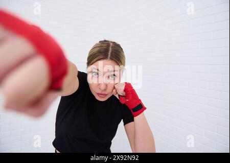 Combattante déterminée avec des bandes rouges sur ses poings, pointant vers l'avant regardant la caméra, isolée sur fond de mur blanc. Jeune femme boxeur 40 s. Banque D'Images