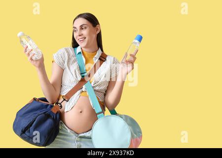 Jeune femme enceinte avec des bouteilles et des sacs d'hôpital de maternité sur fond jaune Banque D'Images