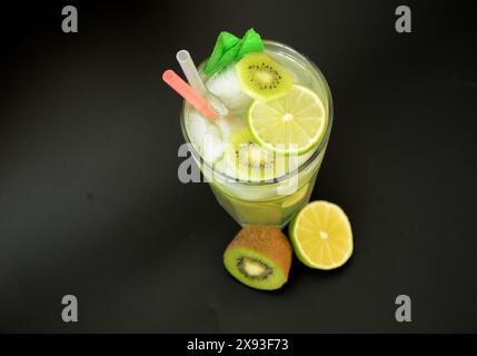 Un verre d'un mélange de jus de fruits avec de la glace et des pailles sur un fond noir, à côté de kiwi mûrs et de citron vert. Gros plan. Banque D'Images