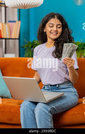 Budget de planification. Riche femme indienne heureuse comptant l'argent comptant utiliser l'ordinateur portable calculer les factures domestiques à la maison. Fille arabe satisfaite des revenus, économise de l'argent pour les vacances planifiées, cadeaux Banque D'Images