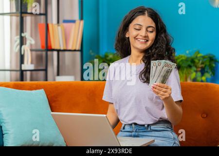 Budget de planification. Riche femme indienne heureuse comptant l'argent comptant utiliser l'ordinateur portable calculer les factures domestiques à la maison. Fille arabe satisfaite des revenus, économise de l'argent pour les vacances planifiées, cadeaux Banque D'Images