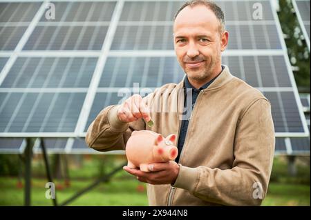 L'homme investit dans des ressources écologiques alternatives. Homme mettant son argent dans une tirelire pour un profit futur. Investisseur dans des vêtements décontractés tenant Moneybox sur fond de panneau solaire. Banque D'Images