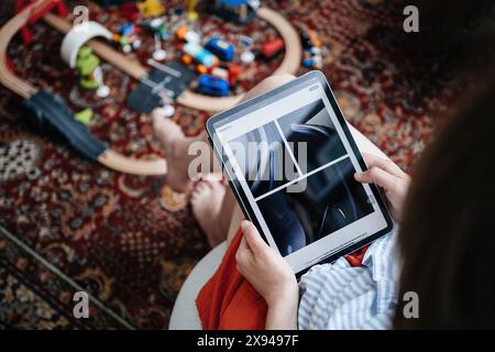 Paris, France - 11 juin 2023 : une femme est assise et navigue sur le site Web d'Apple sur un iPad, visionnant des images détaillées du casque Vision Pro, avec des jouets pour enfants dispersés sur le sol autour d'elle Banque D'Images