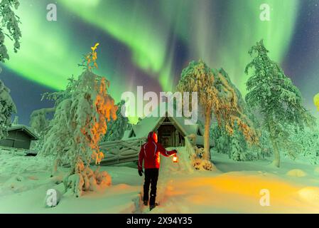 Homme avec une lanterne debout devant une cabane finlandaise typique (kota) sous aurores boréales (aurores boréales), parc national de Pallas-Yllastunturi, Finlande Banque D'Images