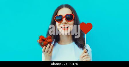 Portrait de belle jeune femme caucasienne avec une poignée de fraises fraîches tenant sucrée portant des lunettes de soleil en forme de coeur rouge sur le goujon bleu Banque D'Images