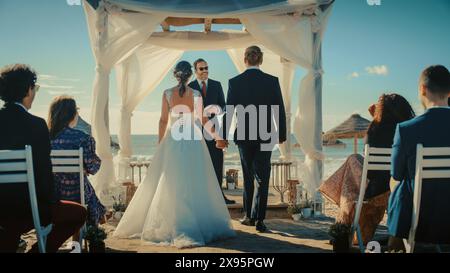 Belle mariée en robe de mariée blanche et beau marié dans un costume noir traditionnel descendant l'allée à un lieu de cérémonie en plein air près de la mer avec des amis multiethniques et divers heureux. Banque D'Images