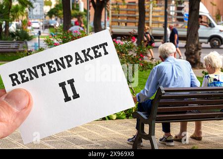 Allemagne - 29 mai 2024 : image symbolique du forfait pension II, Un couple de retraités assis sur un banc de parc tandis qu'une main tient une note avec l'inscription : forfait pension 2. PHOTOMONTAGE *** Symbolbild Rentenpaket II, Ein Rentnerpaar sitzt auf einer Parkbank während eine hand eine Notiz hält mit Aufschrift : Rentenpaket Zwei. FOTOMONTAGE Banque D'Images
