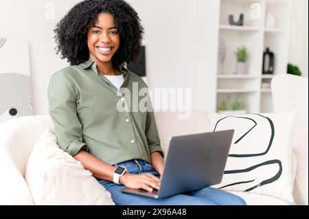 Dans un moment de joyeuse interaction en ligne, une femme afro-américaine sourit en travaillant sur son ordinateur portable dans sa maison lumineuse et moderne. Étudiante étudiante à distance Banque D'Images