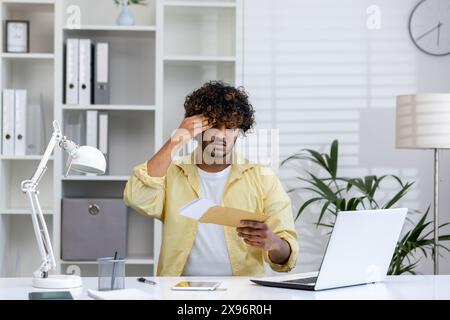 Jeune homme lisant une enveloppe postale et recevant de mauvaises nouvelles dans un bureau à domicile, se sentant stressé et inquiet. Espace de travail moderne avec ordinateur portable et fournitures de bureau. Banque D'Images
