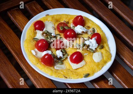 Une omelette garnie de tomates cerises, de fromage blanc et de graines sur une assiette blanche posée sur une table en bois. Banque D'Images