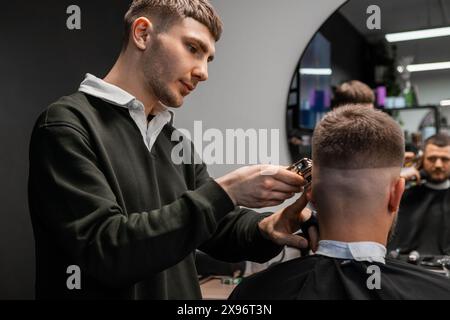 Jeune barbier coupe méticuleusement les cheveux de Mans avec une tondeuse dans le salon de coiffure Banque D'Images